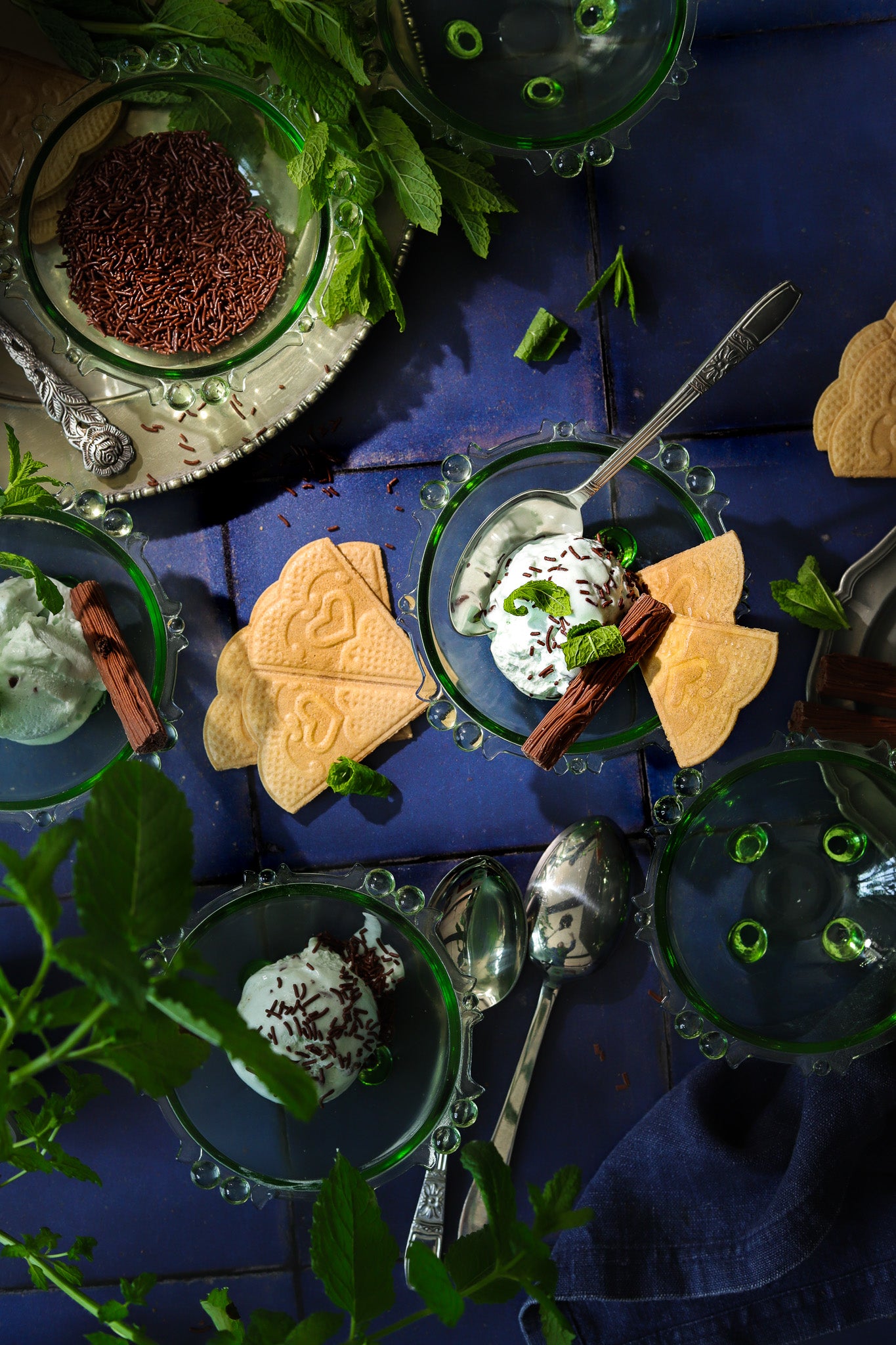 A flat lay of mint chocolate ice creams in vintage green glass dishes styled on a blue tile effect photography backdrop featuring textured grout lines.