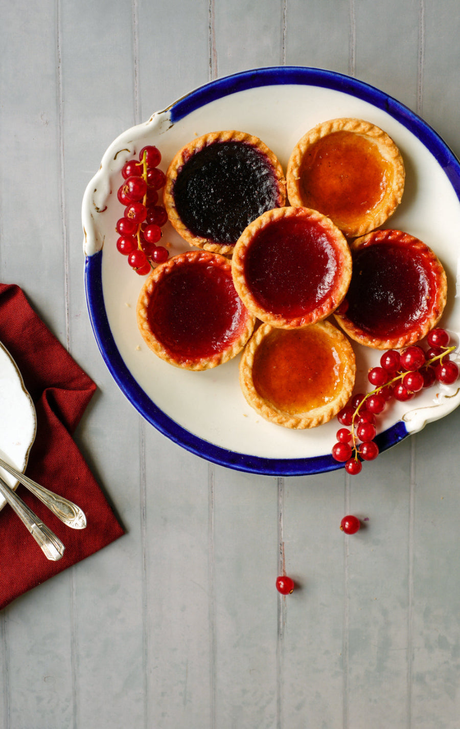 Small jam tarts with different fillings on a vintage plate with redcurrants on a grey painted tongue and groove effect vinyl photography backdrop.