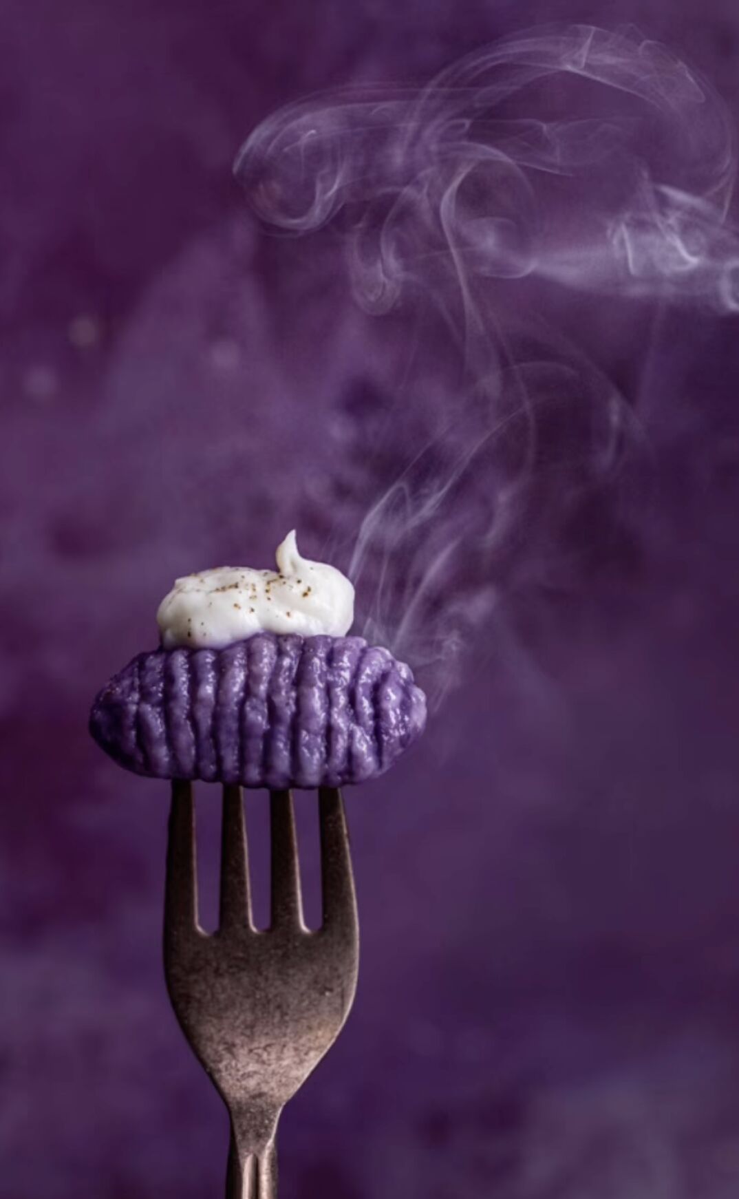 A close-up food photograph of a purple gnocchi topped with a dollop of white cream, held on a copper fork against a purple vinyl food photography backdrop.