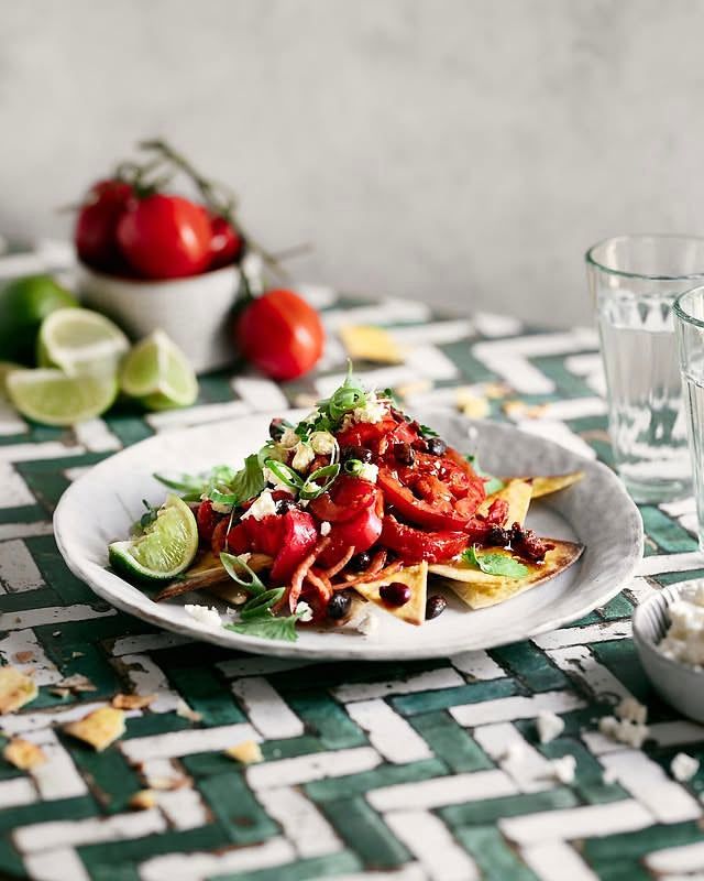 A plated dish of loaded tortilla chips with red tomato salsa, black beans, and fresh cilantro, set on a green and white Moroccan Zellige tile vinyl photography backdrop with limes, tomatoes and glasses in a kitchen setting.