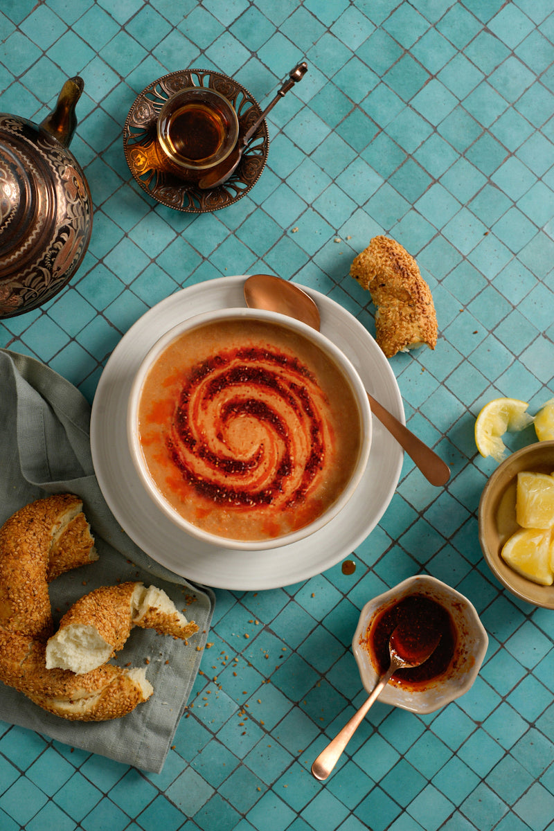 Spicy soup with a spiral pattern on a turquoise tiled table, surrounded by simit and a turkish teacup.