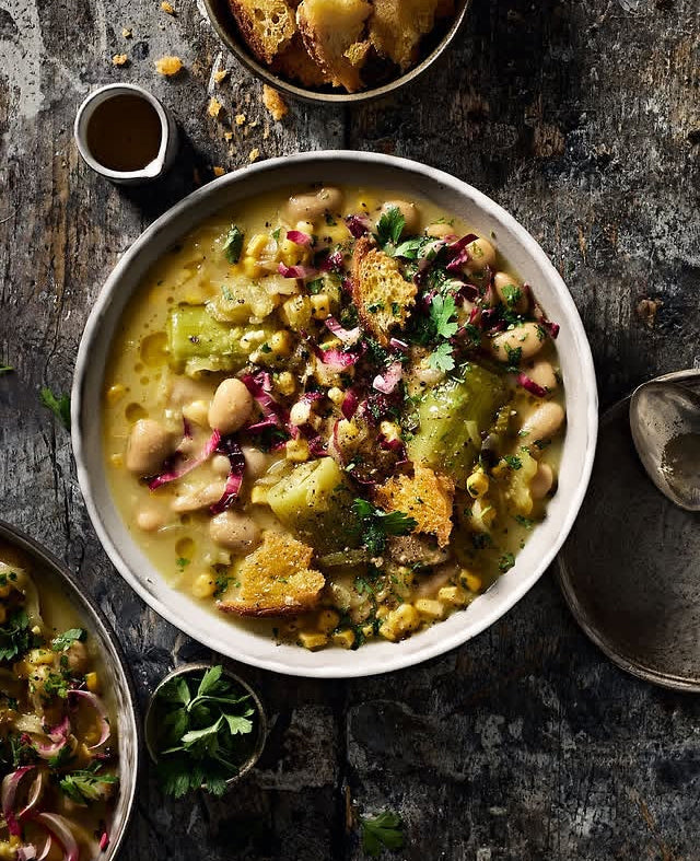 A top-down view of a hearty soup or stew served in a large white bowl, featuring chickpeas, green vegetables, corn, and croutons on a rustic weathered wood effect vinyl photography backdrop.