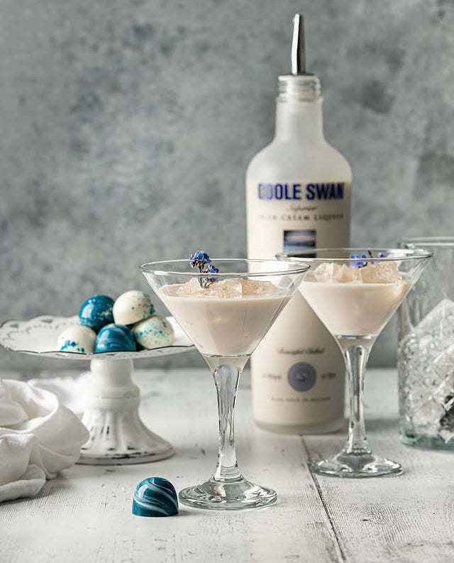 Two chilled martini glasses filled with a creamy pale beverage and garnished with blue flower petals, set on a rustic white vinyl photography backdrop. A frosted bottle labeled Coole Swan cream liqueur stands behind them, with decorative chocolates on a small pedestal and a glass of ice to the side.