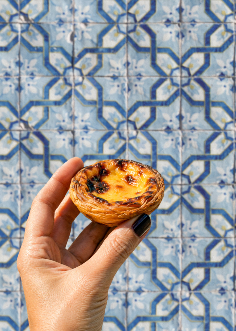 Hand holding a Portuguese pastry against a vinyl photography backdrop featuring a Portuguese azulejo geometric tile pattern in blue, white and yellow.