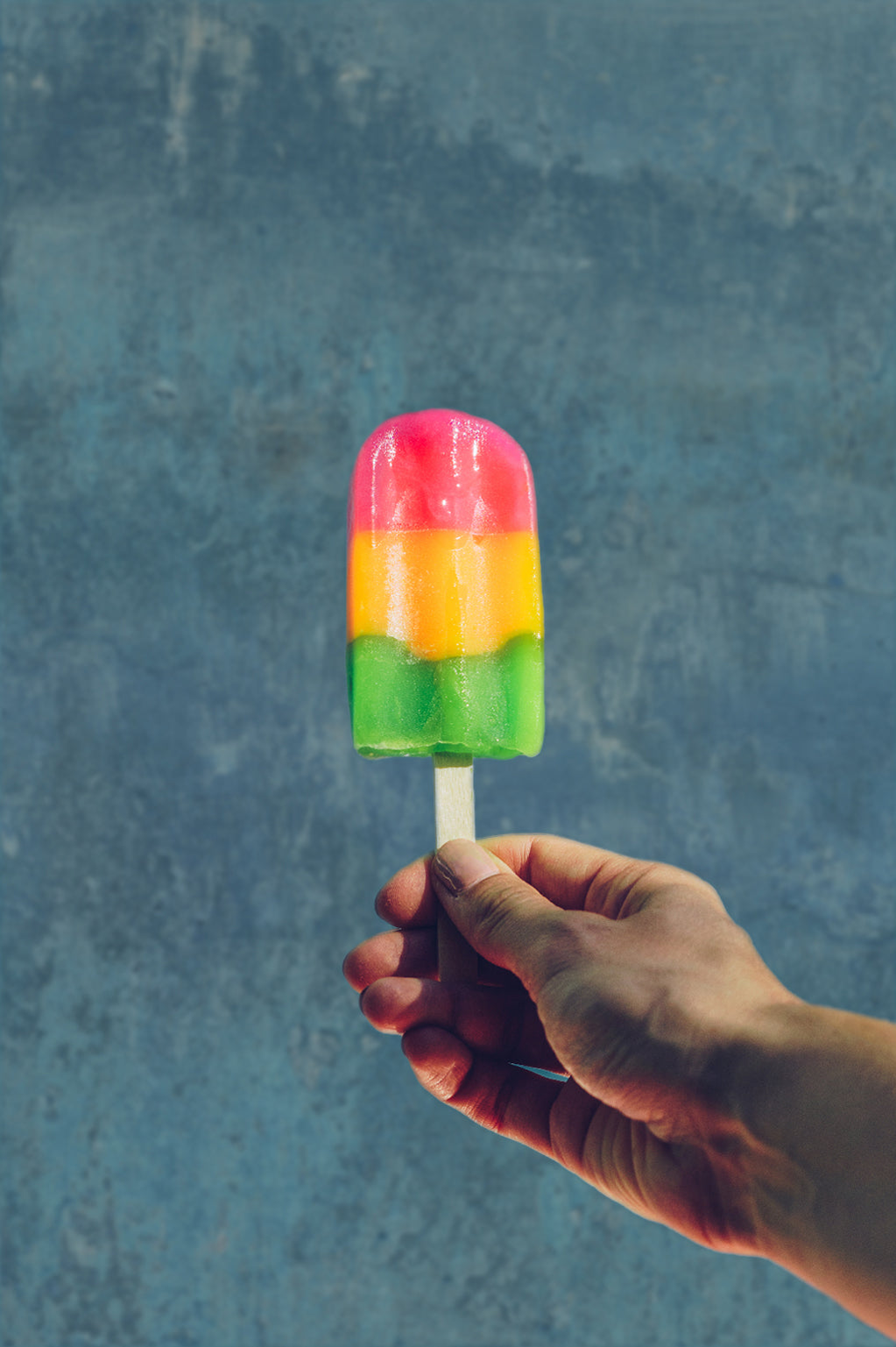 Hand holding a colorful striped popsicle against a blue painted effect vinyl photography backdrop.
