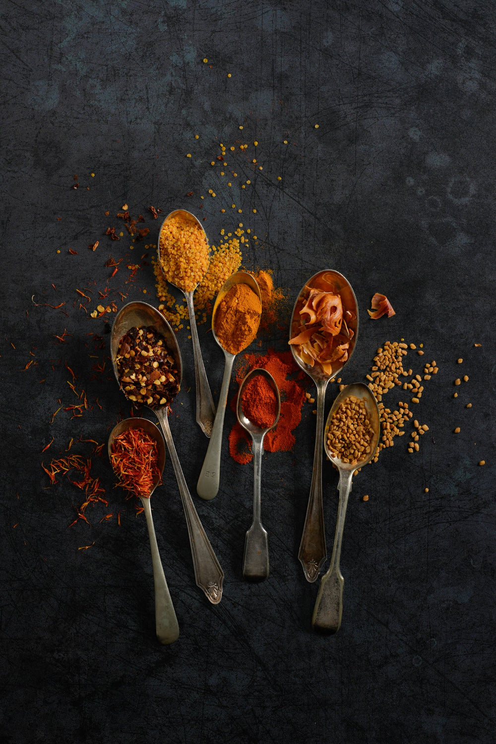 Spoons filled with various spices on a dark grey vinyl photography backdrop with a grungy, rustic pattern.