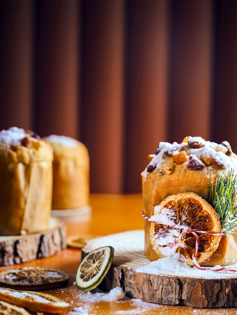 Classic Italian Christmas Panettones with fruits and a slice of orange styled against a tan ribbed vinyl photography backdrop behind.
