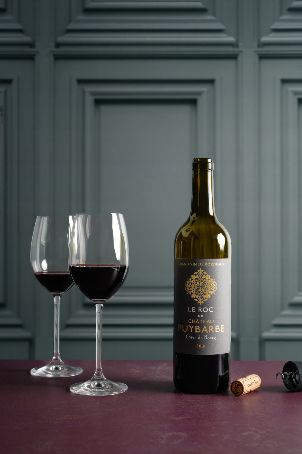 Wine bottle with label, two wine glasses filled with red wine against a sage green vinyl photography backdrop with a wood wall panel design and decorative moulding