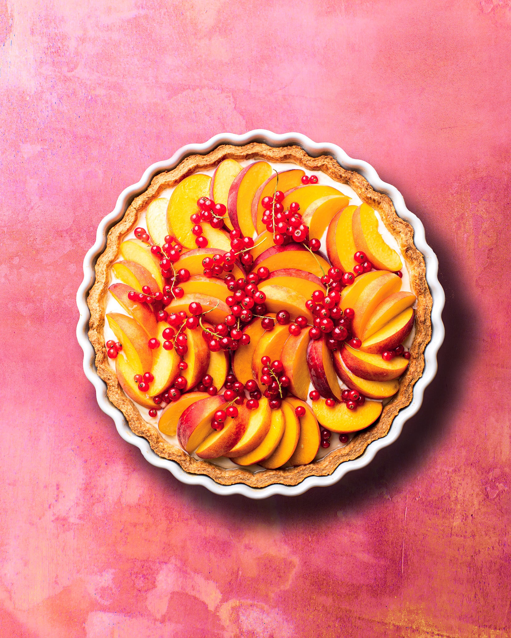 A flat lay of a redcurrant and nectarine flan styled on a pink and peach vinyl photography backdrop with a watercolour effect.