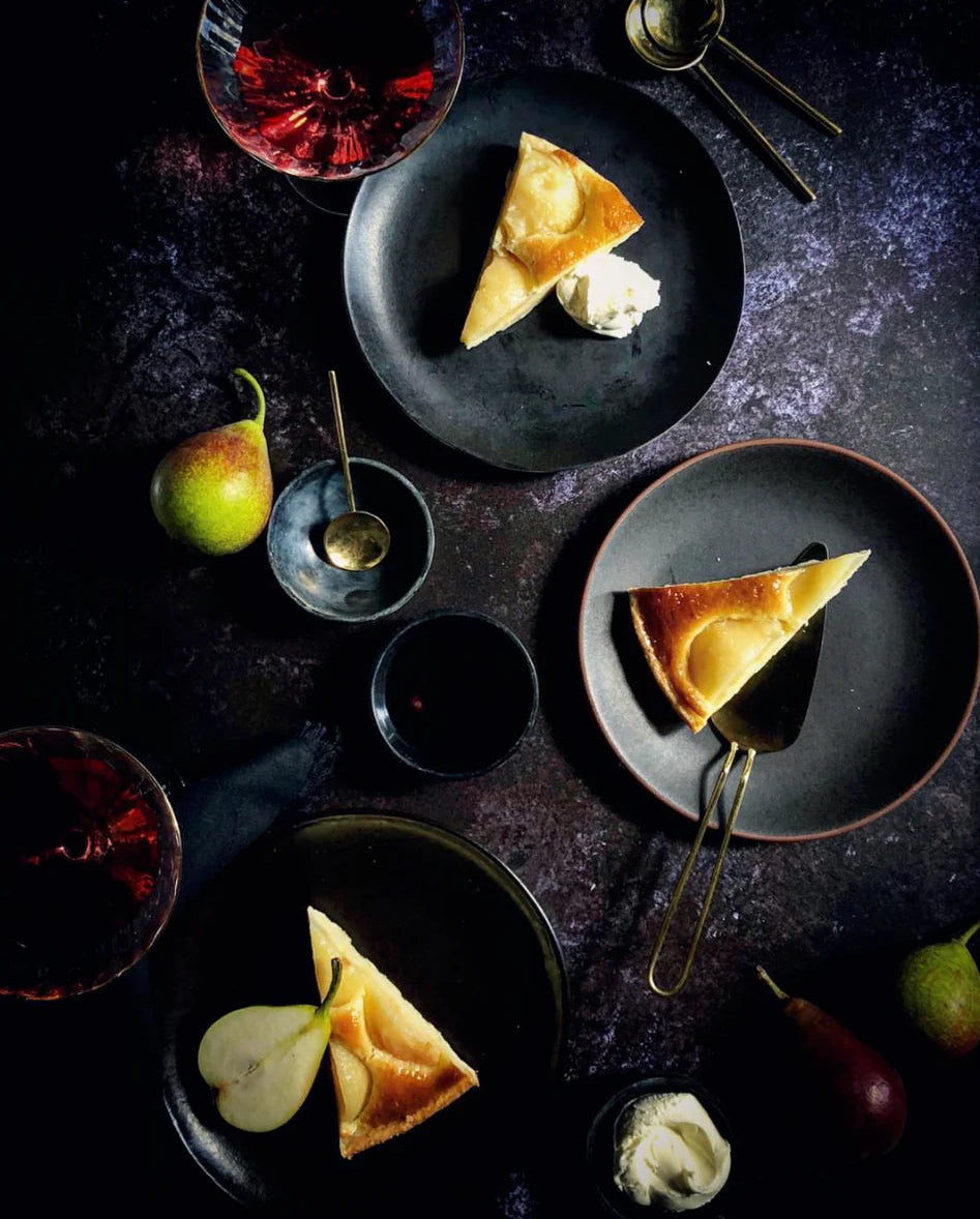 Dessert flat lay scene with slices of cheesecake, pears and red wine styled on a vinyl photography backdrop in mixed dark greys captured from real granite.