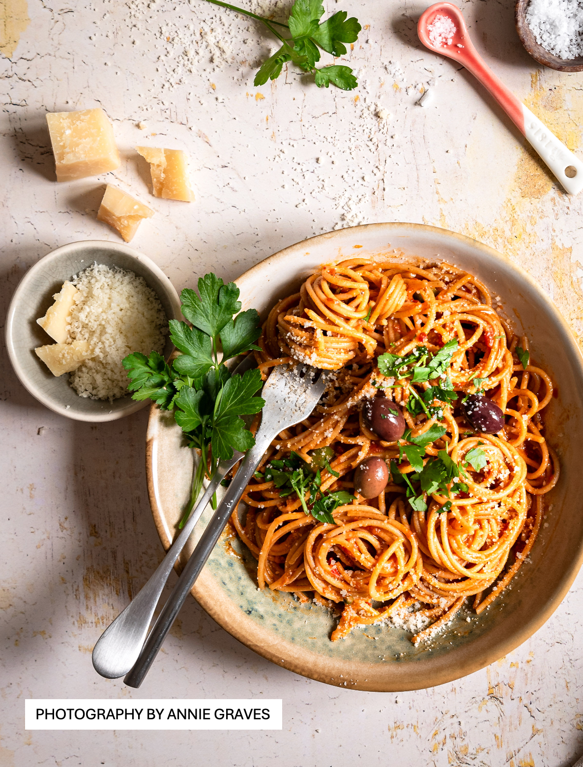 A colourful food photo of a bowl of olive tomato pasta styled on a crackled yellow paint effect photography backdrop for food and product styling.