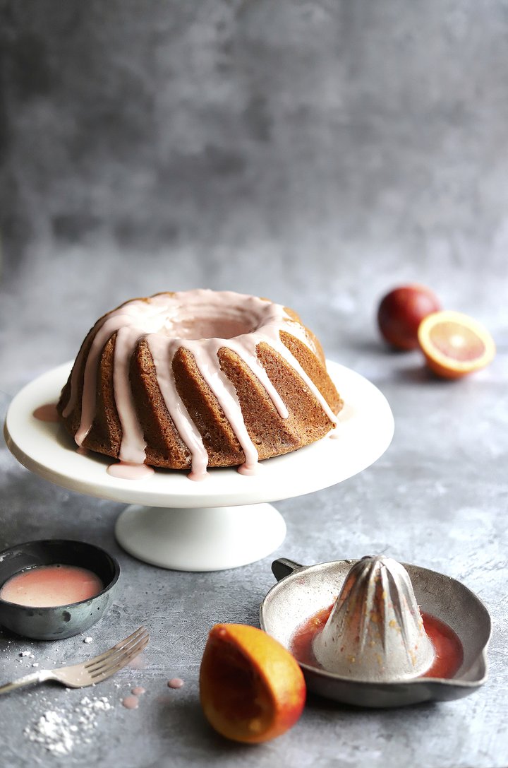 Bundt cake on a white stand with peaches and a juicer on a textured grey plaster effect vinyl photography backdrop.