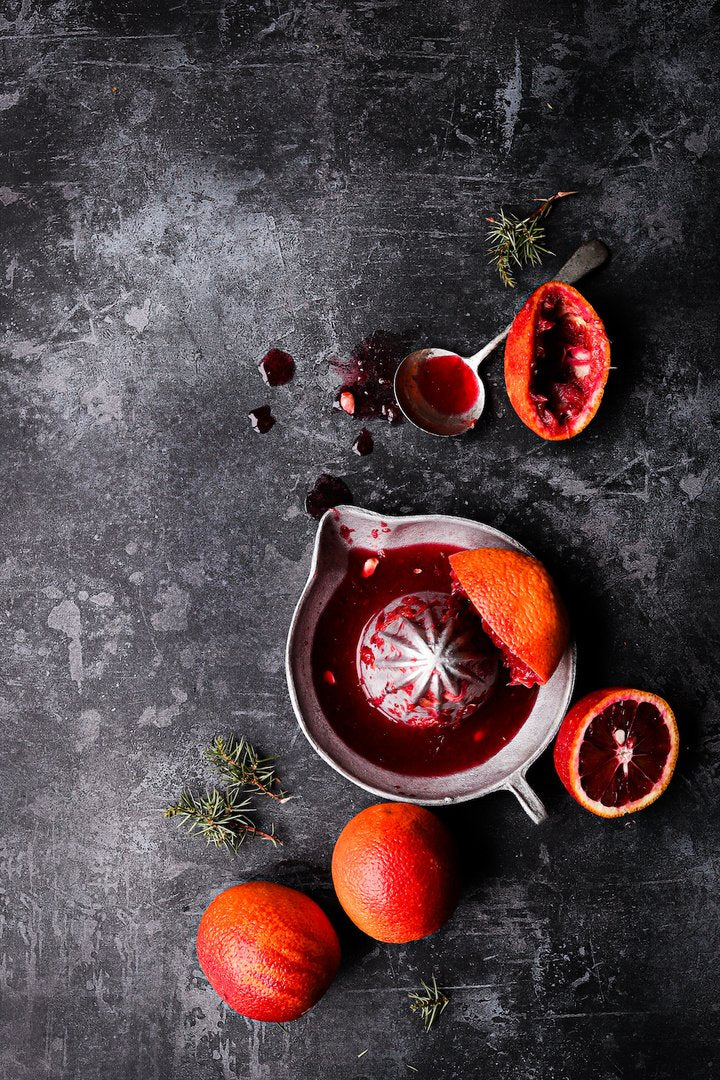 A flat lay of blood oranges in a vintage juicer styled on a grungy grey-black vinyl photography backdrop.