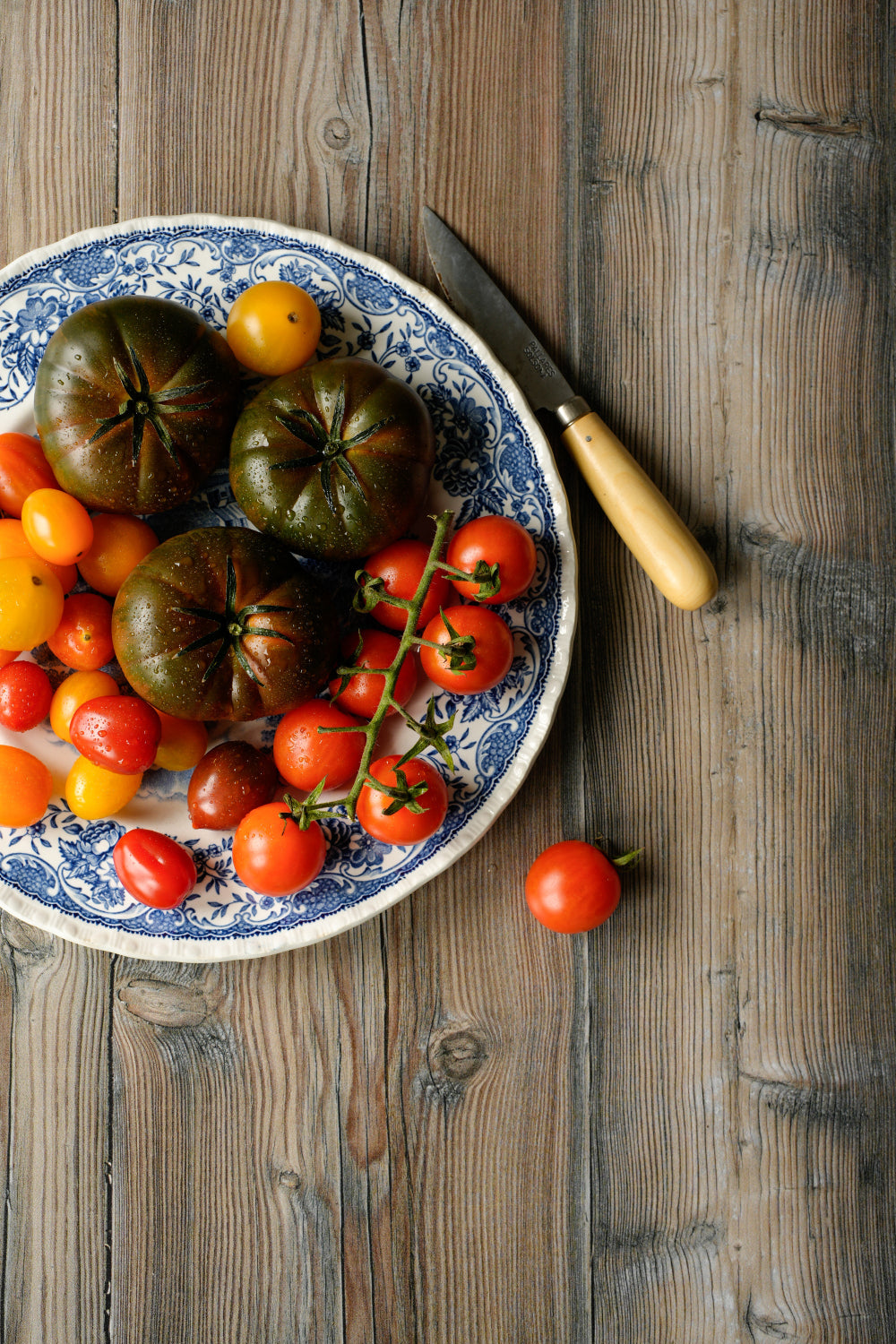 Assorted tomatoes on a decorative plate with a Palleres knife styled on a rustic photography backdrop with a contrasting natural striped woodgrain in beige and browns.
