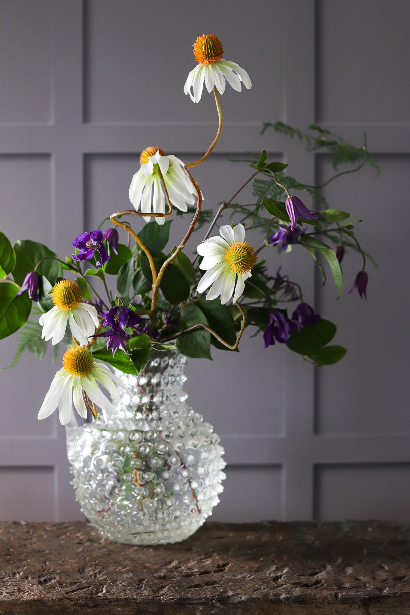 Glass vase and flowers against a panelled wall effect photography backdrop in warm grey with light and shadow details.