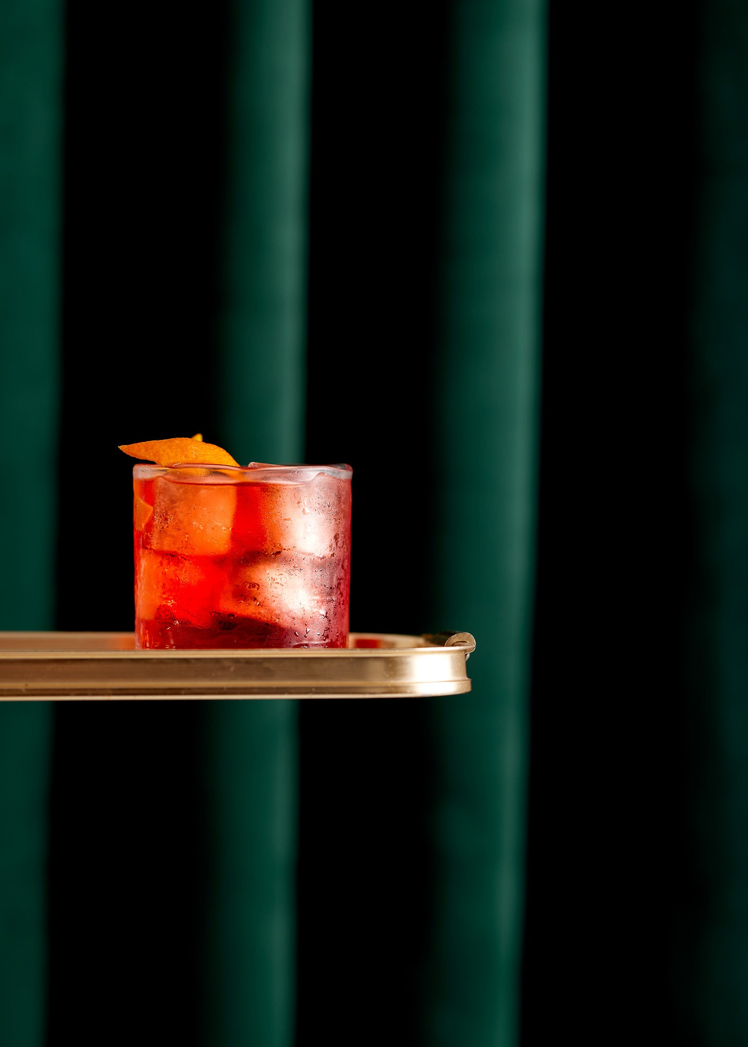 Speakeasy style negroni cocktail on a gold tray against a green velvet curtain-effect vinyl photography backdrop.