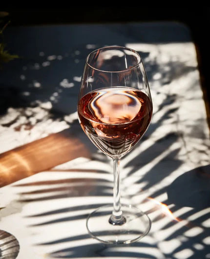 Glass of rose wine on a table with sunlight casting shadows through palm leaves, styled on a beige stone effect vinyl photography backdrop.