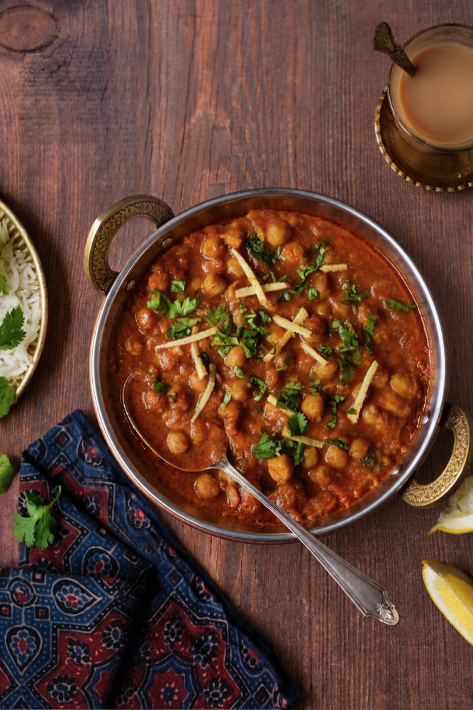 A metal balti dish filled with chana masala garnished with chopped green cilantro and strips of fresh ginger, set on a dark warm wood effect photography backdrop with a spoon, a wood block Indian napkin, and a glass of chai nearby.