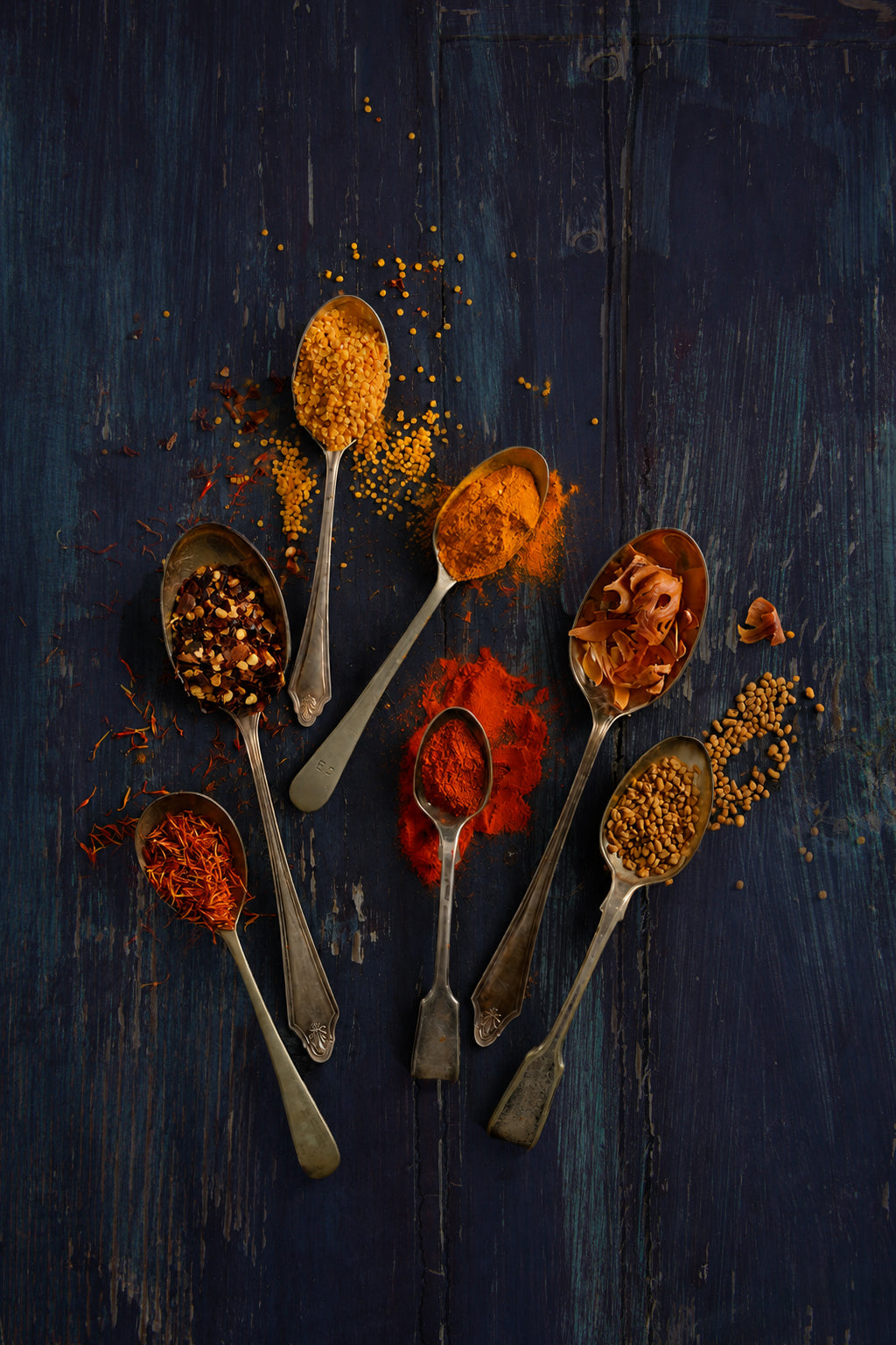 Seven vintage silver spoons with red and orange spices arranged on a wood effect vinyl photography backdrop in indigo and near-black tones, with teal tones showing through. Visible brush strokes, scratches and worn patches give it a rustic, painterly and vintage feel. 