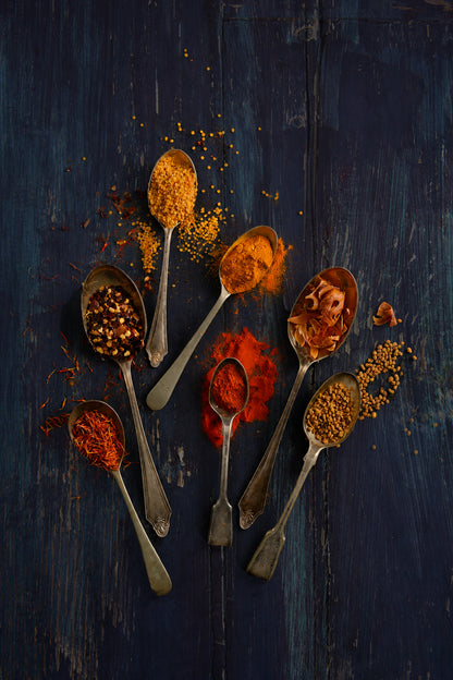 Seven vintage silver spoons with red and orange spices arranged on a wood effect vinyl photography backdrop in indigo and near-black tones, with teal tones showing through. Visible brush strokes, scratches and worn patches give it a rustic, painterly and vintage feel. 