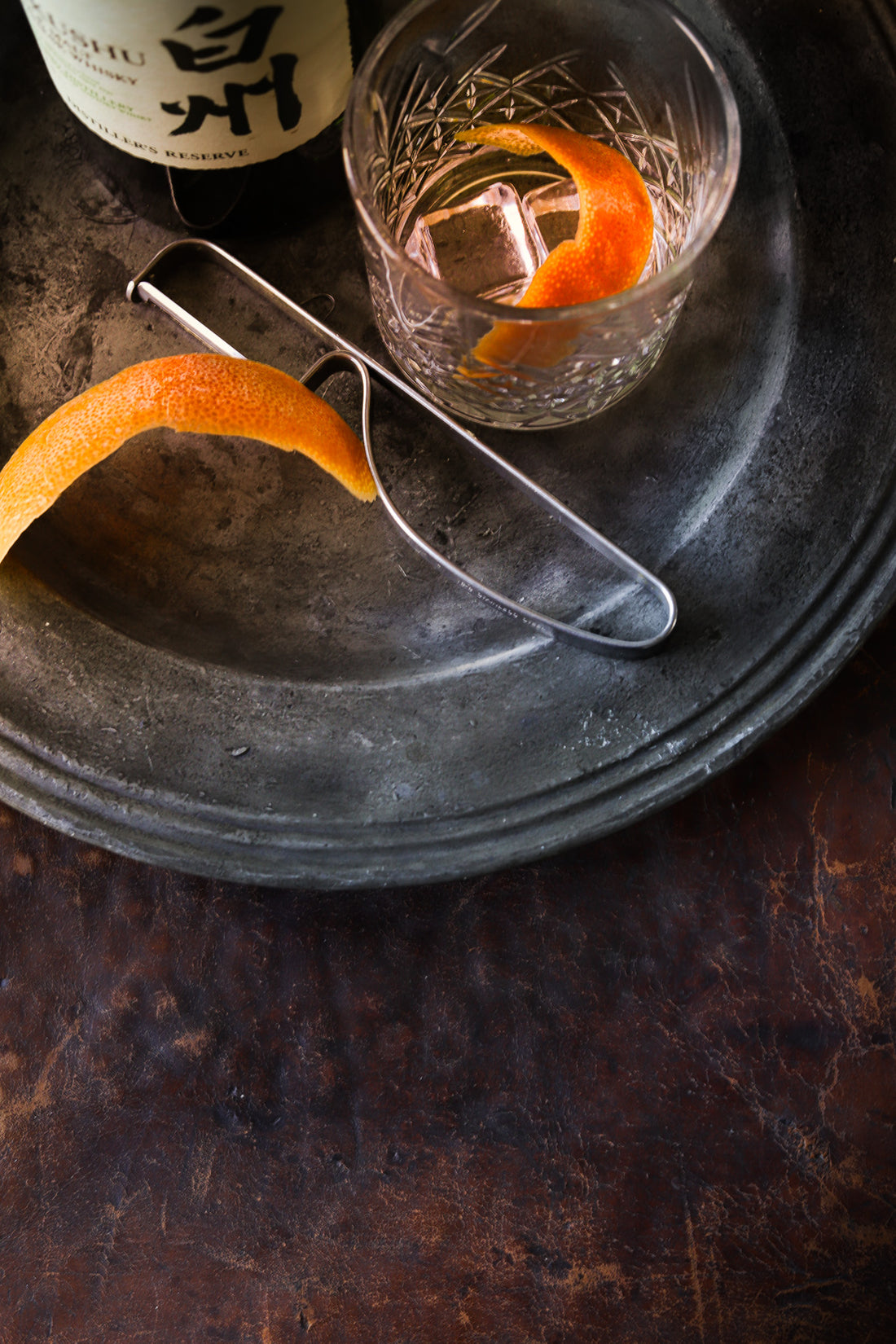 A close-up image pewter plate with a cut crystal glass and edge of a bottle of whisky, styled on a vintage brown leather effect photography backdrop with scratches and other textural details.