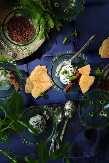 A flat lay of mint chocolate ice creams in vintage green glass dishes styled on a blue tile effect photography backdrop featuring textured grout lines.
