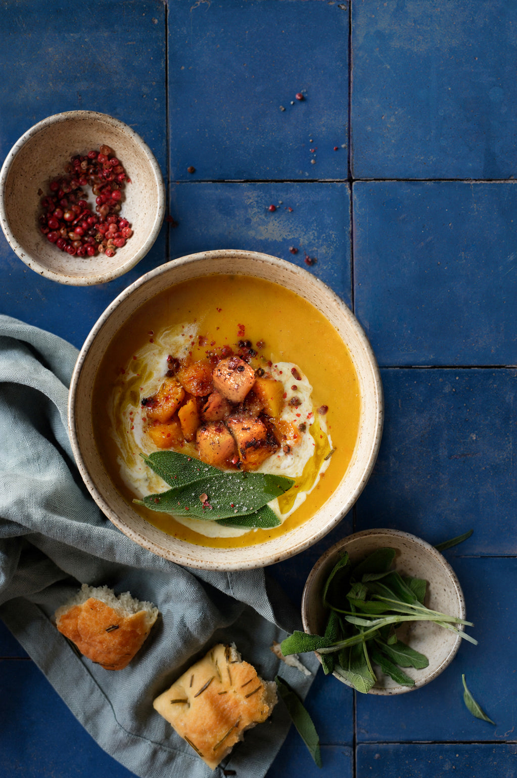 Rustic food flat lay of pumpkin soup, with smaller dishes of fresh sage and pink peppercorns styled on a blue tile effect photography backdrop featuring textured grout lines.