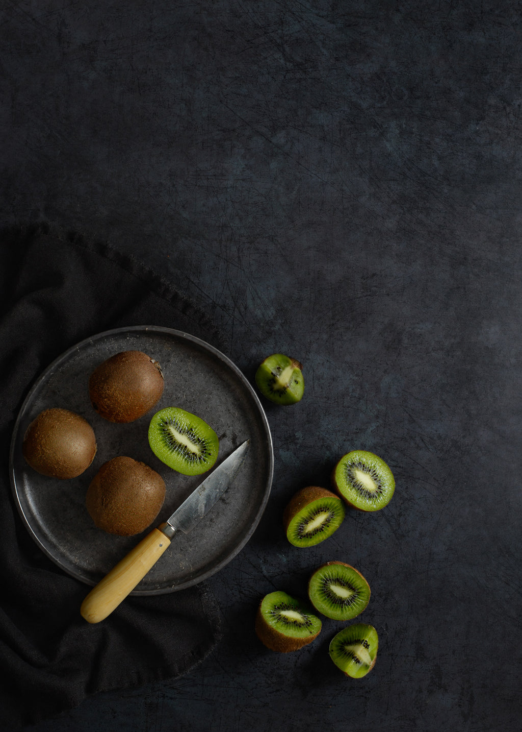 Kiwi fruit arranged on a dark grey metal-effect photography backdrop, with whole kiwis on a round old metal plate, sliced kiwi pieces scattered nearby, and a Palleres knife resting on the plate.