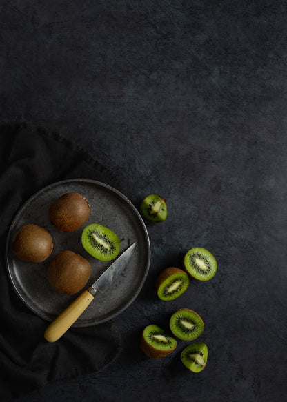 Kiwi fruit arranged on a dark grey metal-effect photography backdrop, with whole kiwis on a round old metal plate, sliced kiwi pieces scattered nearby, and a Palleres knife resting on the plate.