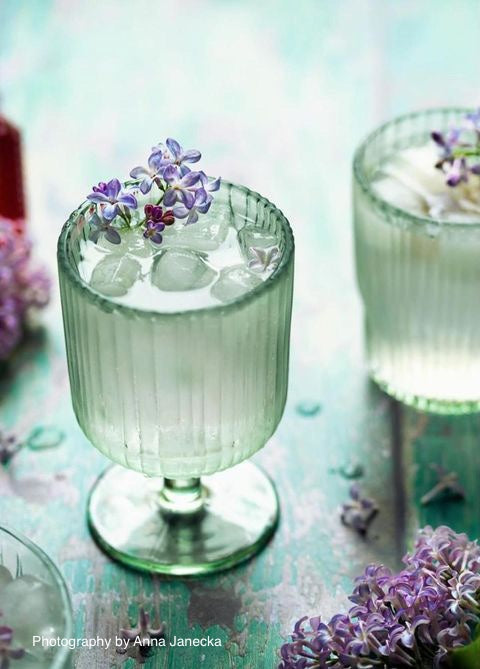 Two green glasses with purple flower buds styled on a green flaky wood plank effect vinyl photography backdrop with a textured surface.