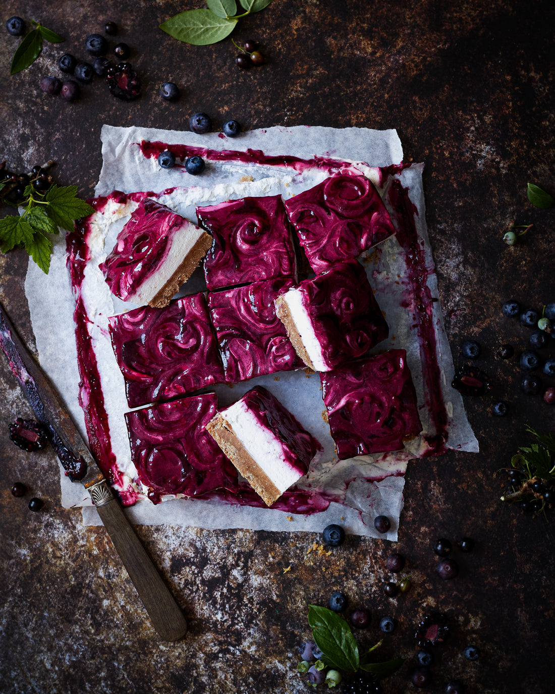 Flat lay of handmade blueberry cheesecake styled on a mottled brown vinyl photography backdrop with dark, umber tones and delicate rusted patches.
