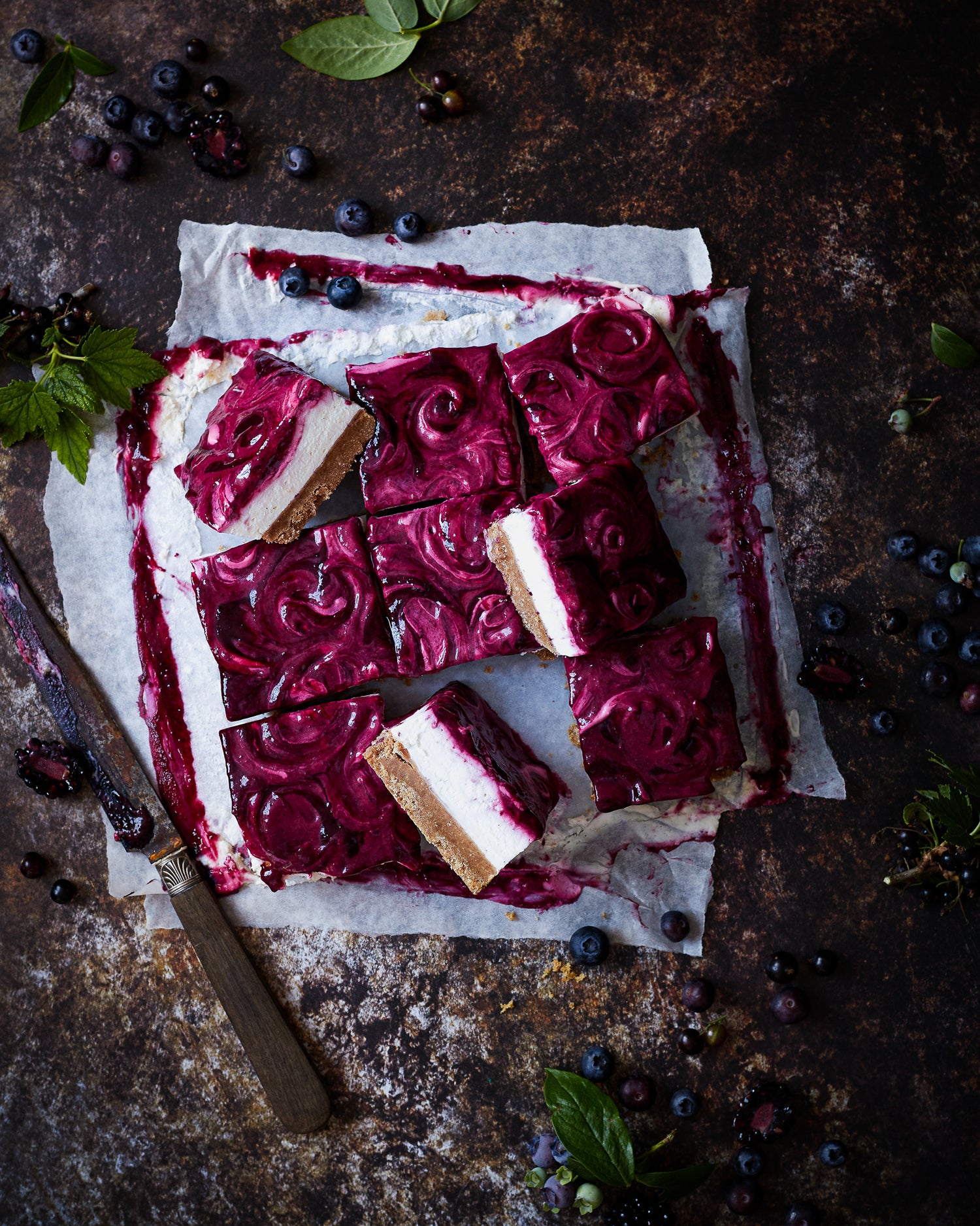 Flat lay of handmade blueberry cheesecake styled on a mottled brown vinyl photography backdrop with dark, umber tones and delicate rusted patches.