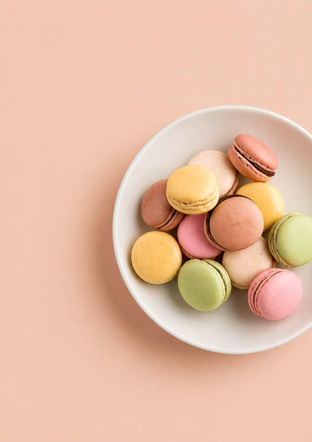 Assorted colorful macarons in a white bowl on a pale peach background