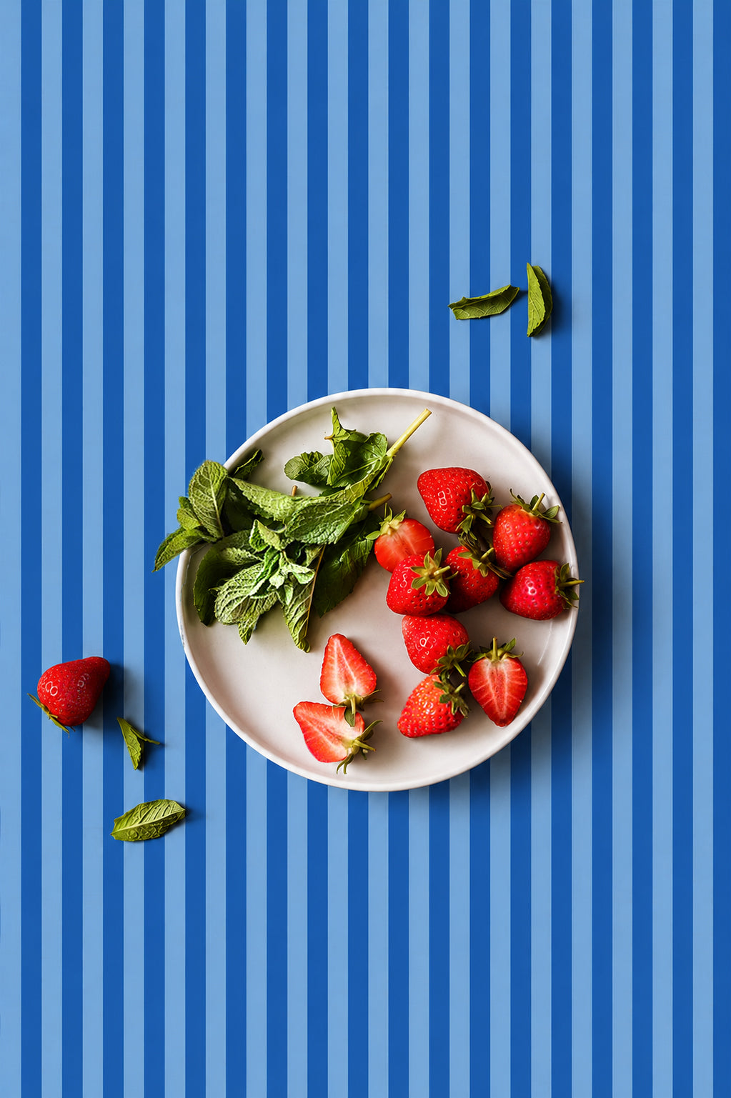 A white plate filled with fresh red strawberries and green mint leaves, arranged on a vertical blue striped waterproof vinyl photography backdrop designed for food styling and product photography.