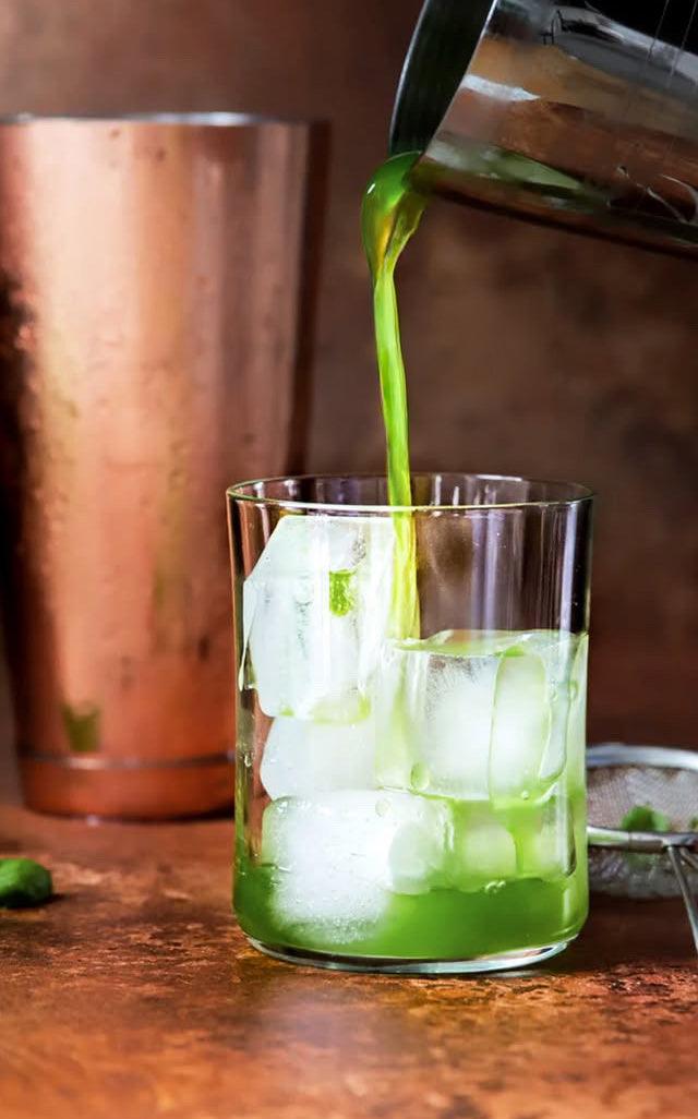Green liquid being poured into a glass with ice cubes on a copper effect vinyl food and product photography backdrop 