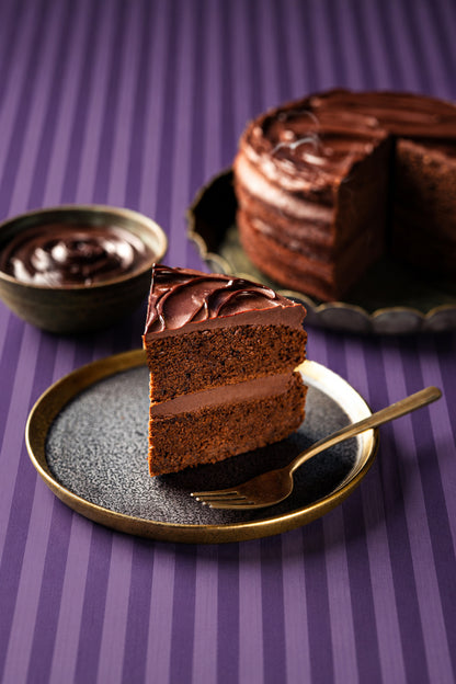 A slice of chocolate cake with thick chocolate frosting on a dark plate with a gold rim, alongside a small bowl of chocolate sauce. The background shows a larger chocolate cake on a matching plate, all set against a purple striped vinyl photography backdrop.
