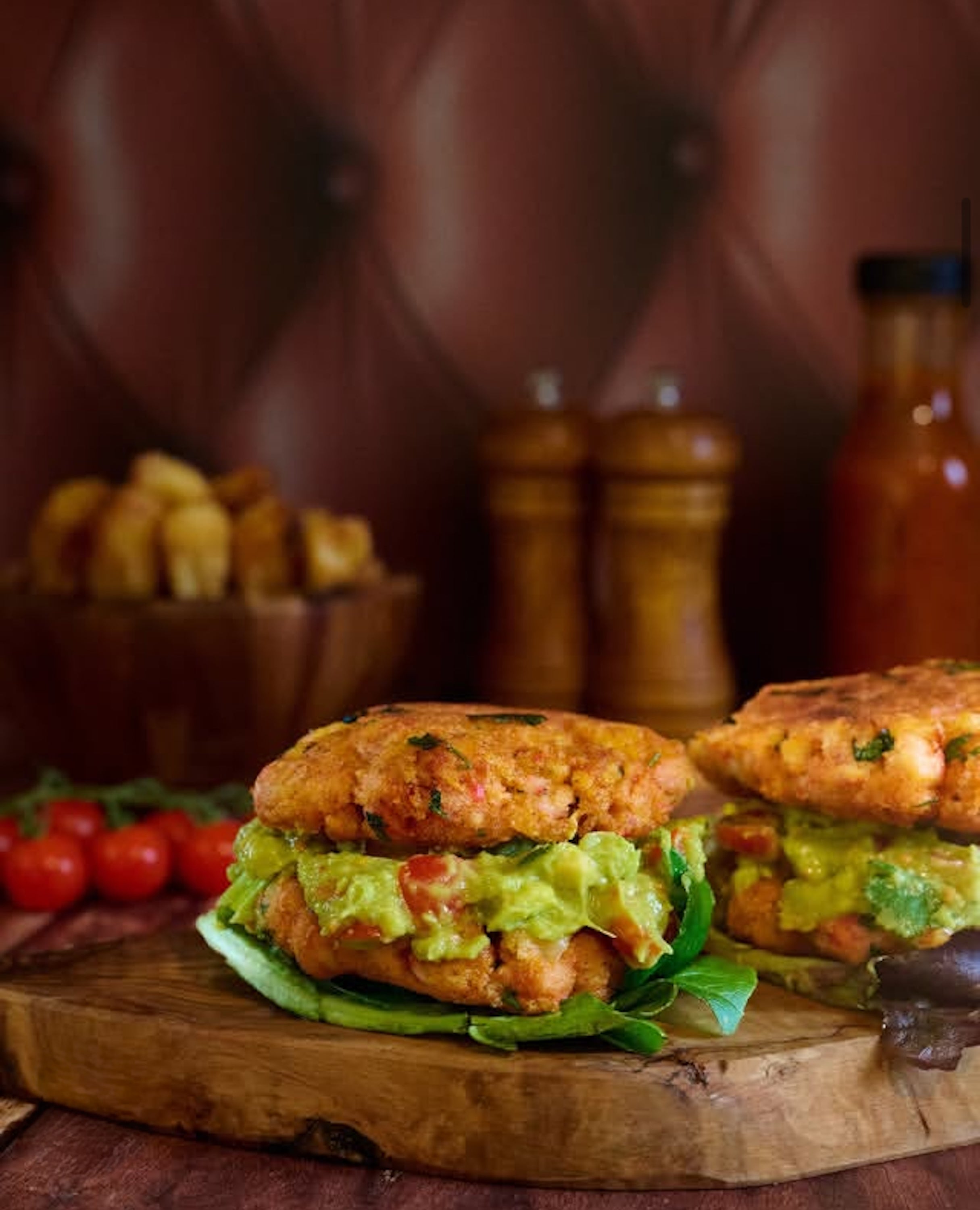 Two salmon burgers with guacamole and lettuce against a tan leather effect photography backdrop with button details, resembling a Chesterfield sofa.