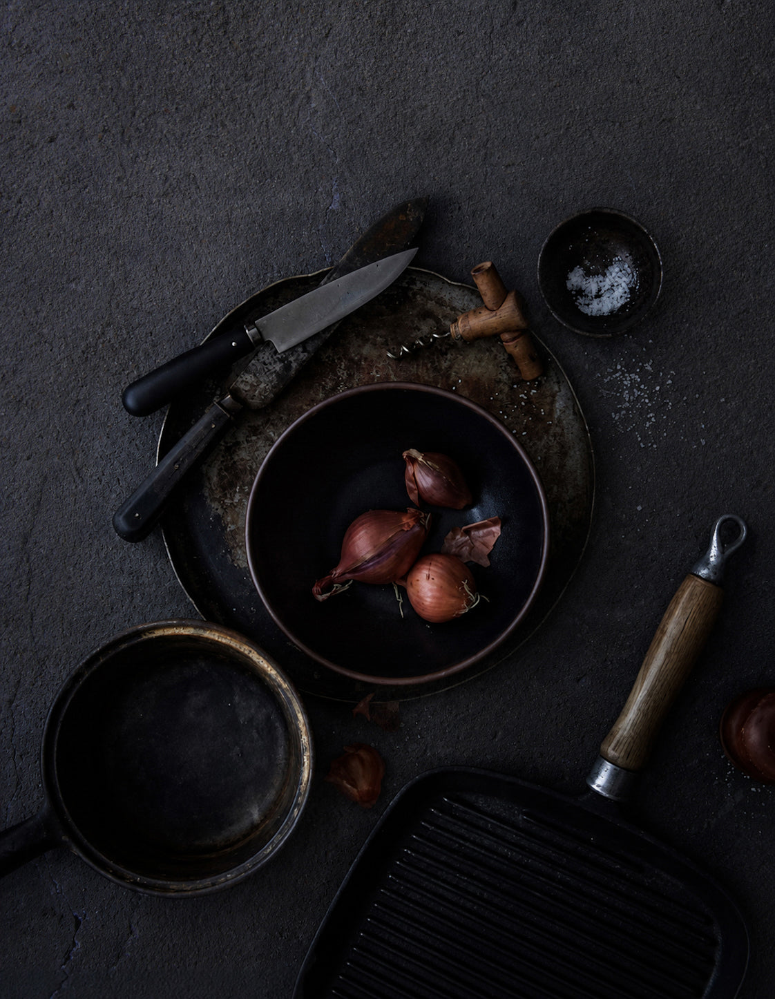 A dark, moody flat-lay of cooking ingredients and utensils on a textured plaster effect dark vinyl photography backdrop for food and product styling.