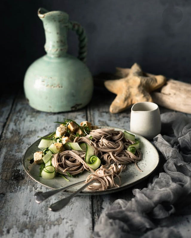 A styled food scene showing a plate of soba noodles with cucumber, green onions set on a rustic wood effect vinyl photography backdrop with a ceramic pitcher, starfish and grey fabric.