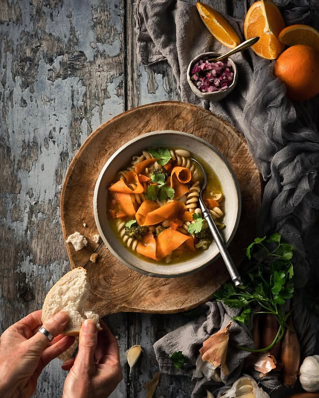 A bowl of vegetable pasta soup with orange carrot slices, green herbs styled on a wood board with bread, garlic, oranges, and a small bowl of red onion on a rustic grey wood effect vinyl photography backdrop. Hands are visible holding a piece of bread.