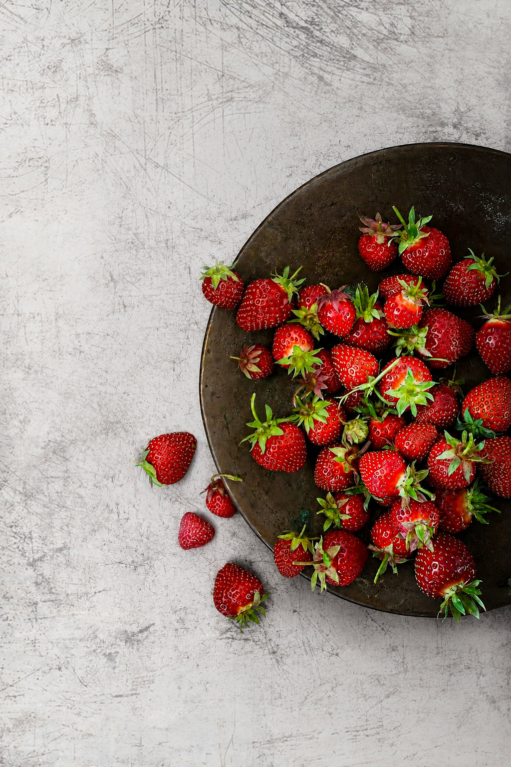 Fresh strawberries on a dark metallic plate shot on an abstract weathered white photography backdrop with grey scratches and a metal patina.