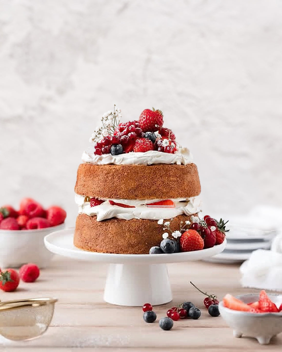 A two-tier sponge cake with layers of whipped cream and fresh berries, displayed on a white cake stand on a wooden table with a white vinyl photography backdrop behind the set.