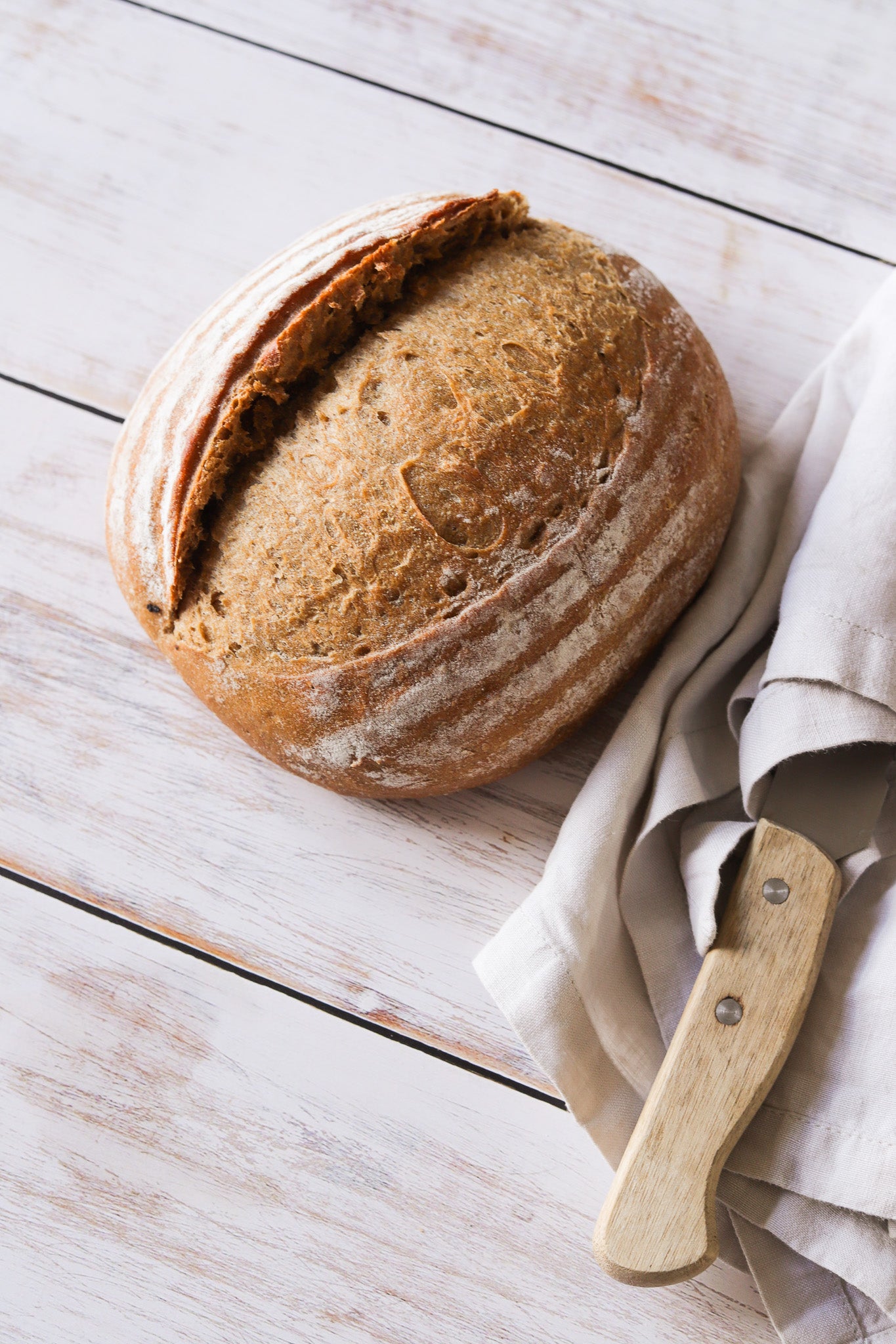 A artisan loaf of bread with a vintage knife, styled on a white and grey wood plank effect photography backdrop.