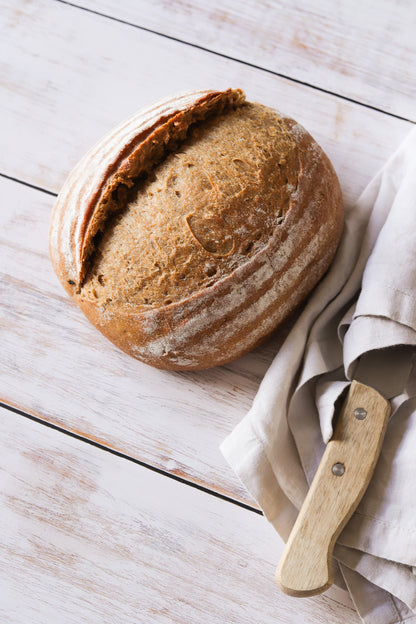 A artisan loaf of bread with a vintage knife, styled on a white and grey wood plank effect photography backdrop.