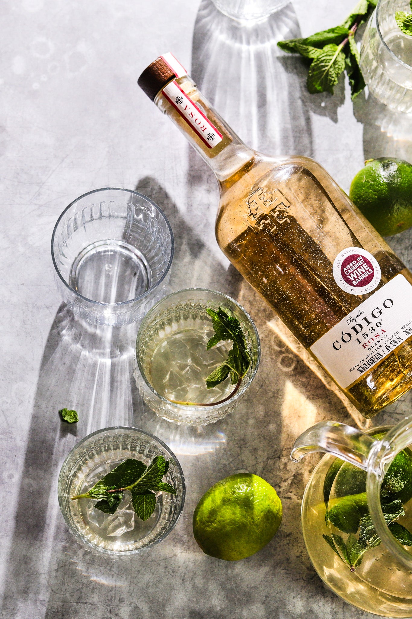 Flat lay of a bottle of tequila with mojito cocktails styled on a weathered grey metal effect vinyl photography backdrop.