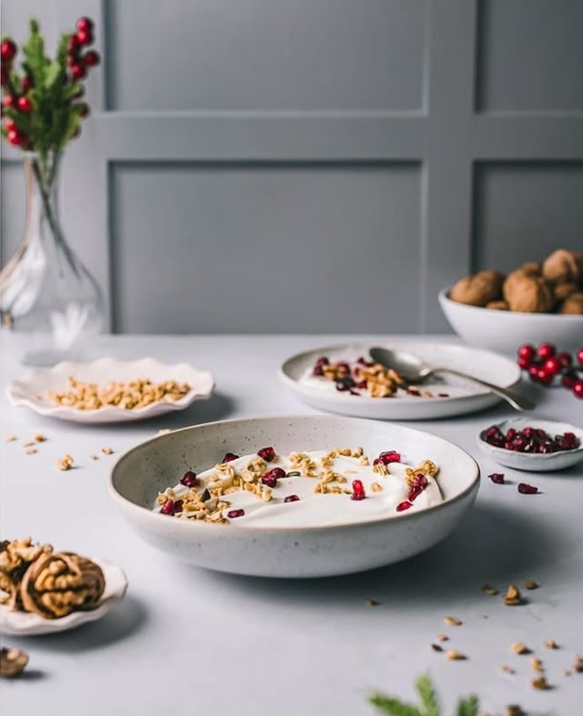 Bowl of yogurt with granola and berries on a table with a gray wall and decorative items in the background.