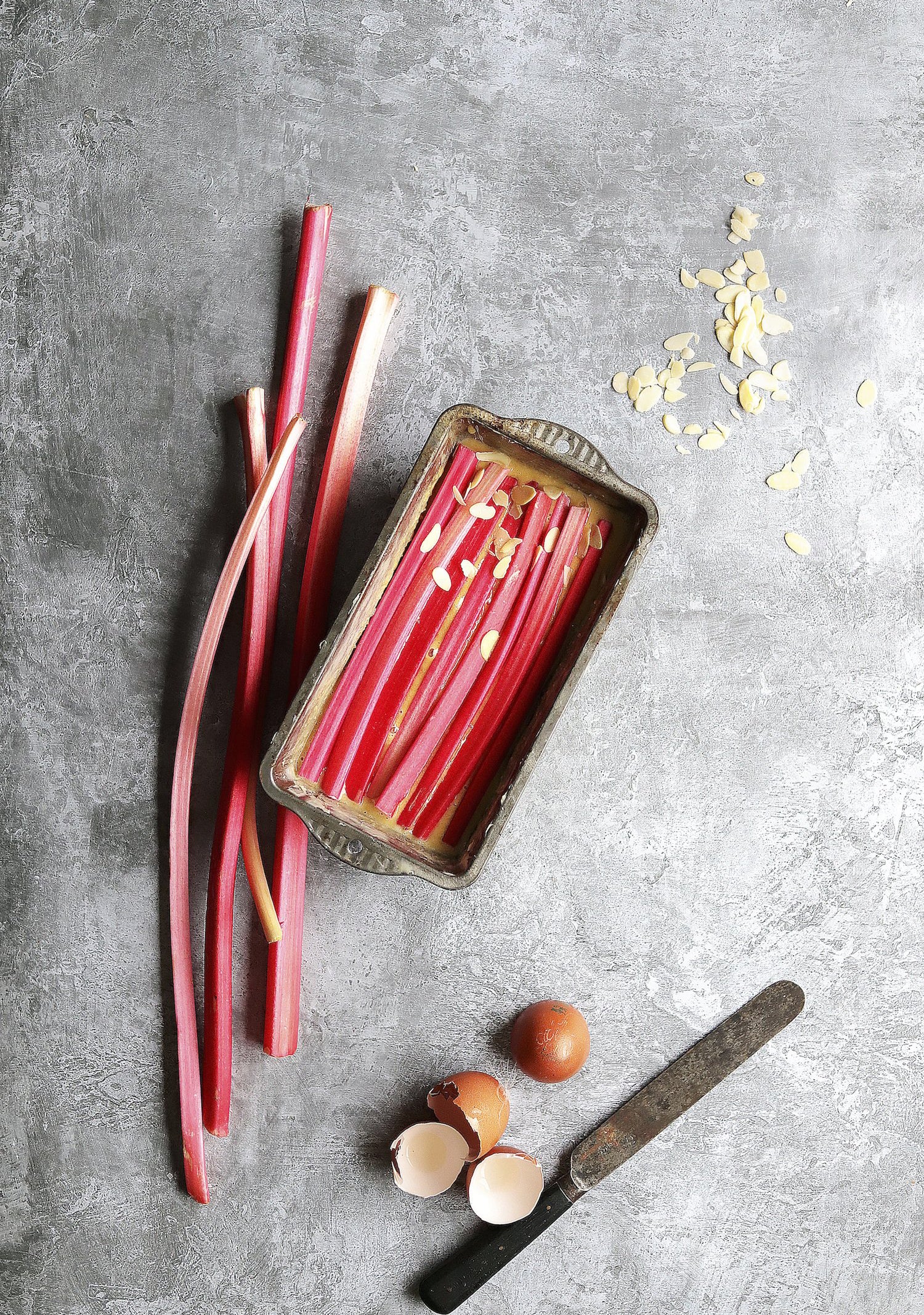 Red rhubarb stalks in a metal container on a gray surface with a knife and eggs.