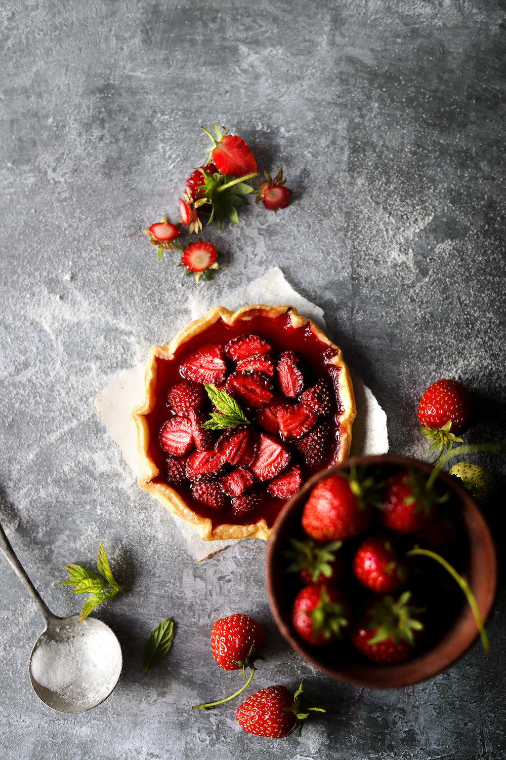 Flat lay of a strawberry flan and cut fresh strawberries styled on a textured grey plaster effect vinyl photography backdrop.