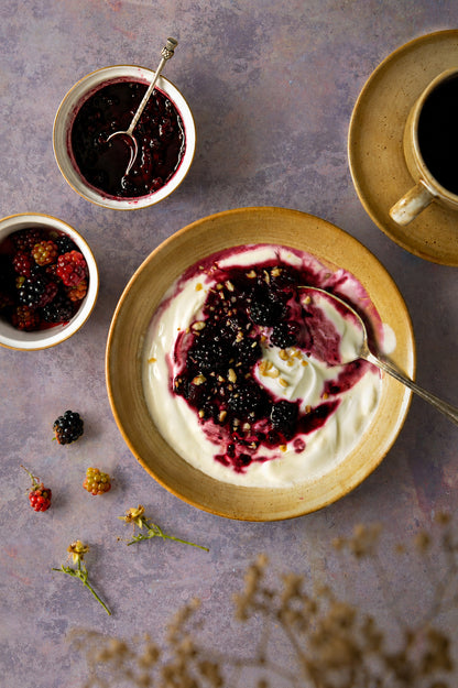 A flat lay of blackberry yoghut and black coffee in French vintage crockery, styled on a light purple and copper effect vinyl photography backdrop.