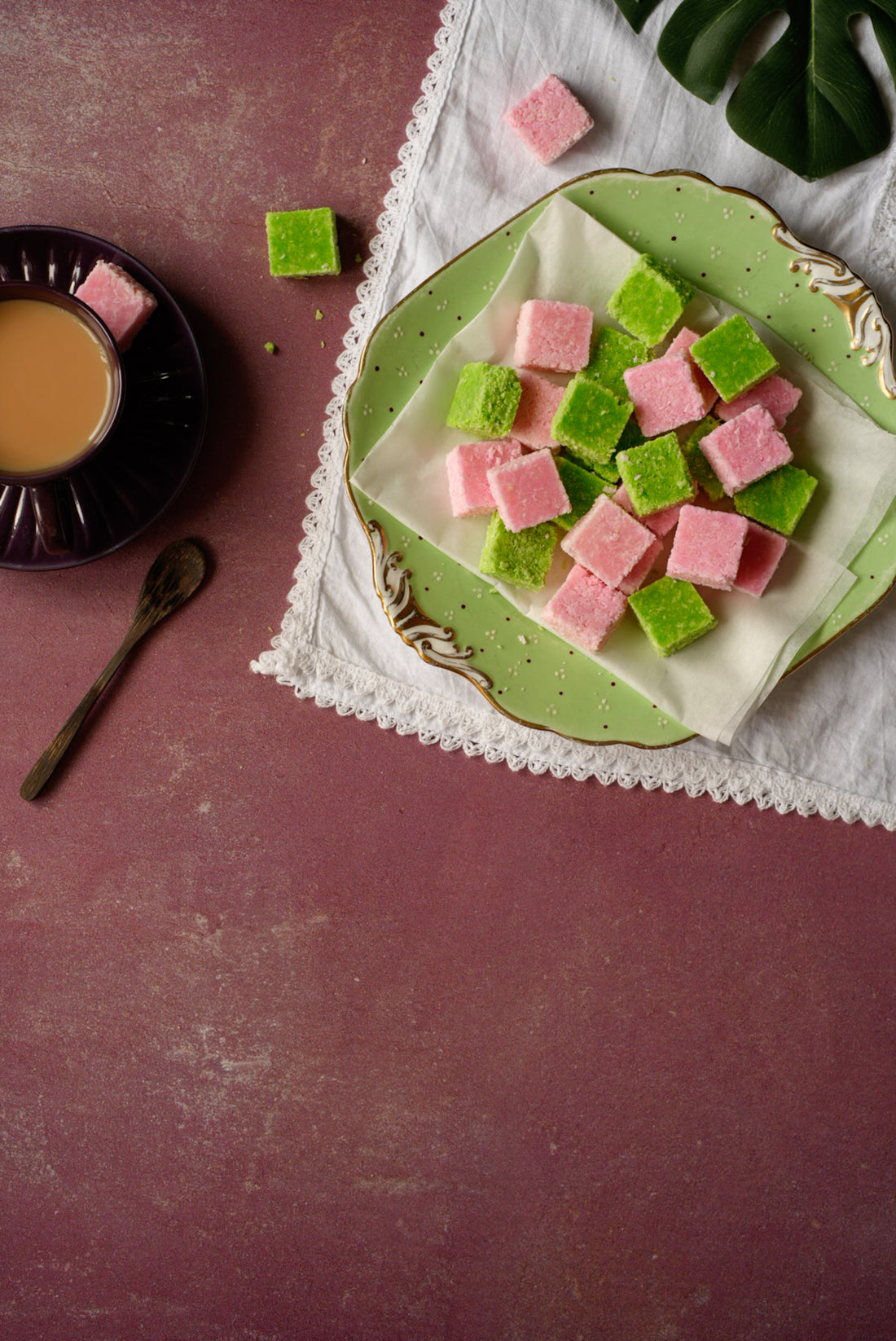 Green and pink square-shaped coconut toffee on a decorative plate with a cup of coffee and spoon on a maroon 